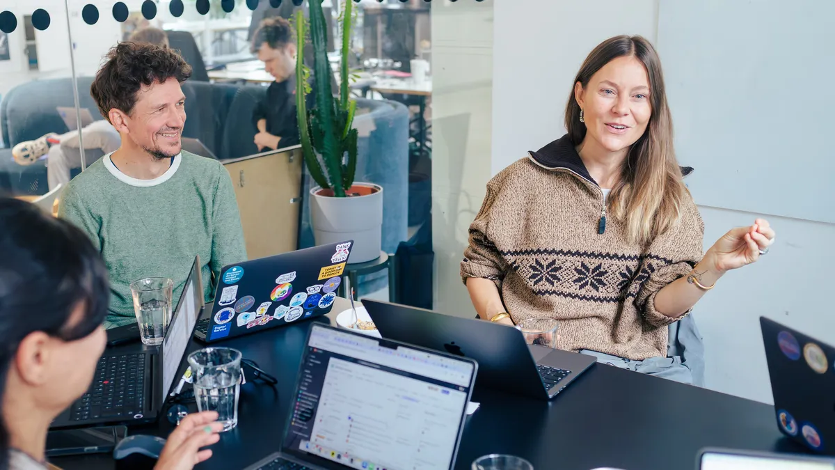 Drei Mitarbeitende des DigitalService sitzen an einem Tisch und sprechen miteinander. Vor ihnen stehen geöffnete Laptops. Eine Mitarbeitende rechts im Bild spricht lächelnd und gestikuliert mit dem Arm. Die anderen Mitarbeitenden hören ihr zu.