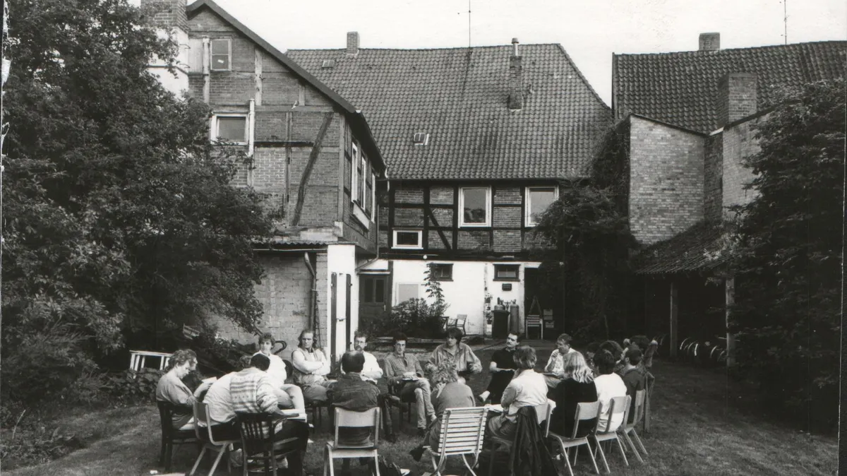schwarz-weiß Aufnahme von 17 Menschen in einem engen Kreis aus Stühlen im Hof vor einem historischen Fachwerkhaus, links und Rechts Bäume, im Hintergrund Fahrräder