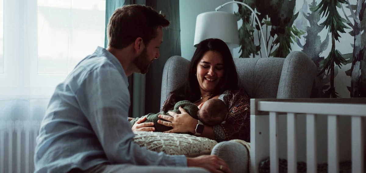 Ein Vater und eine Mutter in einem Kinderzimmer. Die Mutter hält ein Baby im Arm. Man sieht das Kind von hinten. 