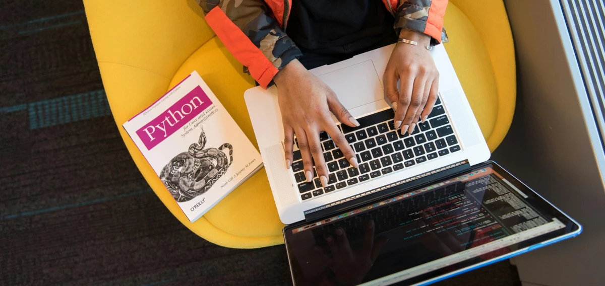 Eine Person sitzt auf einem gelben Sessel, ein Macbook auf dem Schoß und ein Buch zu „Python" neben sich. 