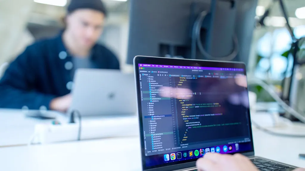 Ein Laptop steht auf einem Schreibtisch. Hinter diesem Laptop ist eine Person unscharf zu erkennen. Auf dem Display des Laptops befindet sich Code.