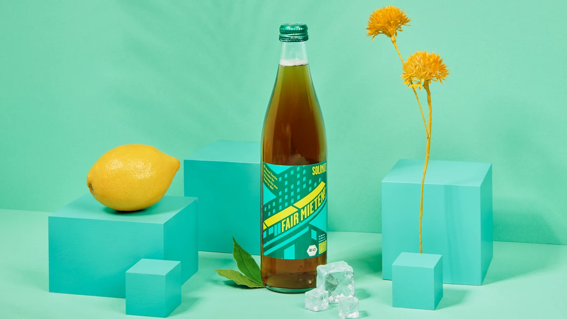 Mate-Flasche steht inszeniert zwischen einer Zitrone und einer Zierpflanze vor türkisem Hintergrund.