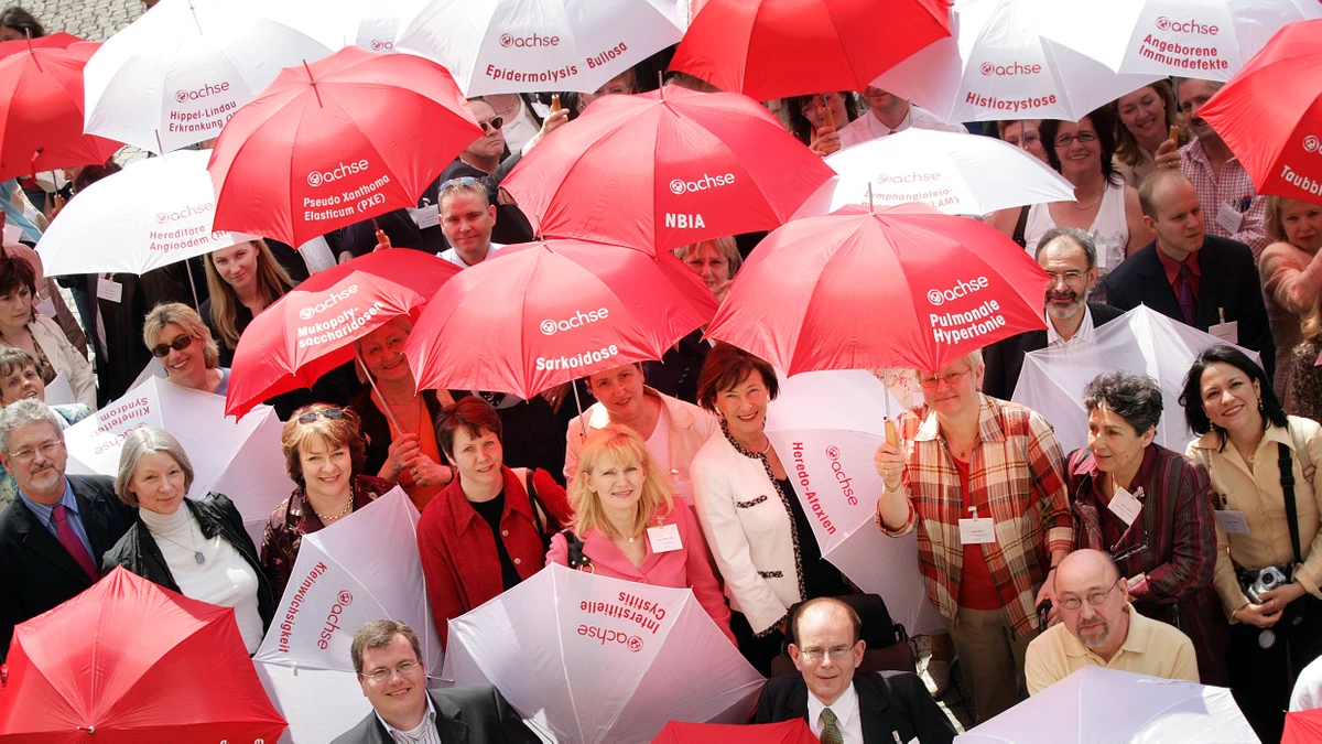 Diverse Personengruppe angeschnitten hält rote und weiße Schirme mit dem Logo der ACHSE in die Kamera. Versinnbildlicht die Vielfalt der Erkrankungen unter dem Dach der ACHSE.