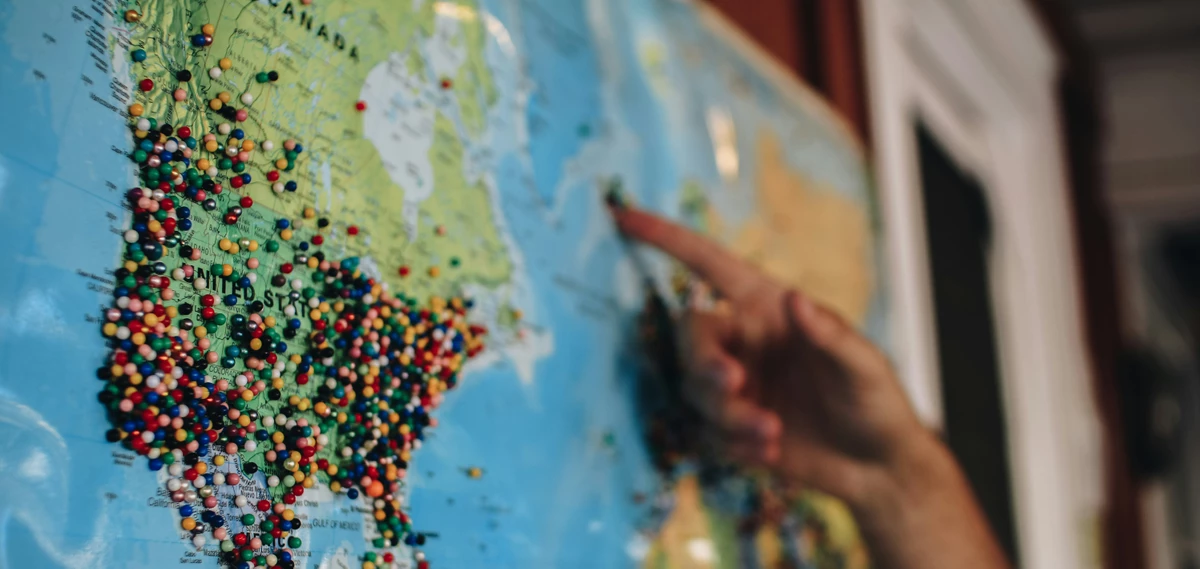 Eine Weltkarte mit zahlreichen bunten Stecknadeln, die verschiedene Standorte markieren. Im Vordergrund zeigt eine Hand auf einen bestimmten Punkt. Das Bild symbolisiert die globale Präsenz und Vernetzung von NGOs (Nichtregierungsorganisationen) und NPOs (Non-Profit-Organisationen).