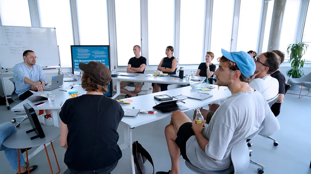 ine Gruppe von 10 Personen in informeller Kleidung, die in einem modernen, lichtdurchfluteten Büro um einen Tisch herum sitzen – mit einem Whiteboard und einem Poster des Servicestandards hinter ihnen.