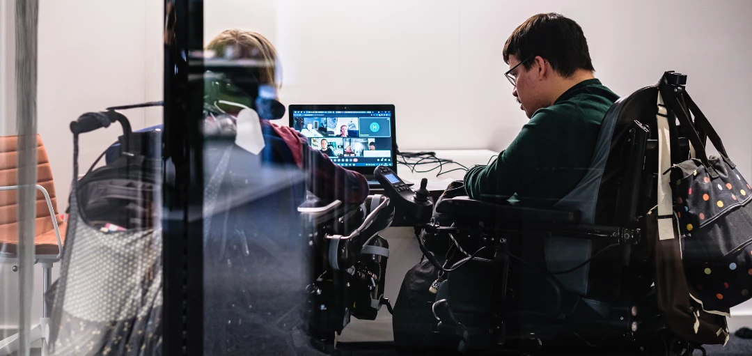 Zwei Menschen im Rollstuhl sitzen am Schreibtisch in einem Online-Meeting 