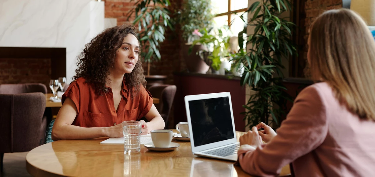 Zwei Frauen sitzen an einem runden Coffee Table in einem Jobinteriew in einem Loft oder Café. Die Interviewering hat einen Laptop vor sich. Vor beiden steht eine Tasse. 