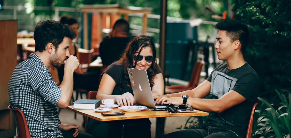 Drei junge Menschen sitzen im Freien an einem Tisch mit einem Laptop und Kaffee und arbeiten gemeinsam an einem Projekt – symbolisch für ein kollaboratives Trainee Programm in einer entspannten Arbeitsatmosphäre.