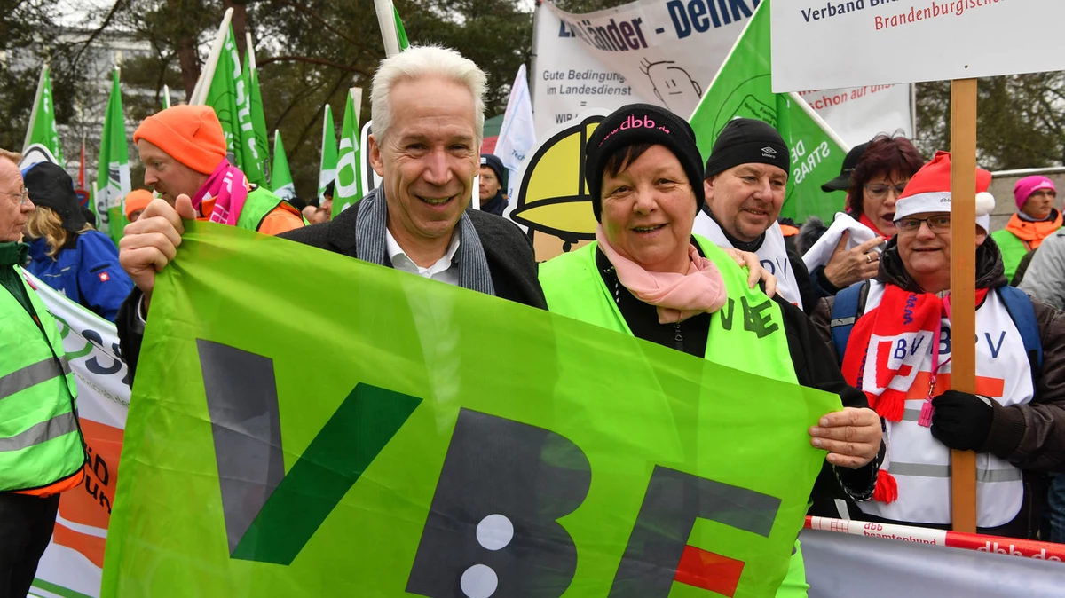 Es sind zwei Personen auf einer Demonstration zu sehen. Im Vordergrund halten zwei Personen eine Fahne, auf der "VBE" steht. Im Hintergrund sind weitere Personen zu sehen, die Fahnen und Banner halten.