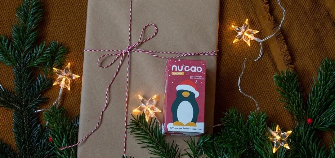 nu+cao Schokoladen-Pinguin auf einem Geschenk mit Tannenzweigen und Sternenlichterkette 