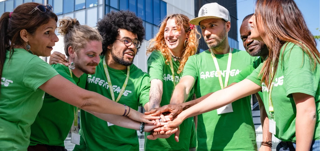Diverses Greenpeace-Team aus sieben Personen steht im Kreis und hält die Hände aufeinander. 