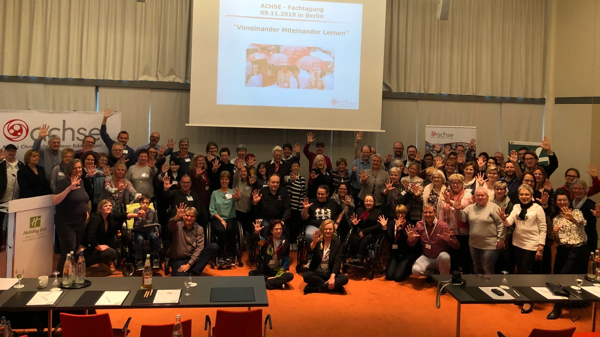 Etwa 70 Personen von schräg oben fotografiert in einem Kongresssaal. Tische im Vordergrund, im Hintergrund  eine Powerpoint sowie Banner mit dem Logo der ACHSE