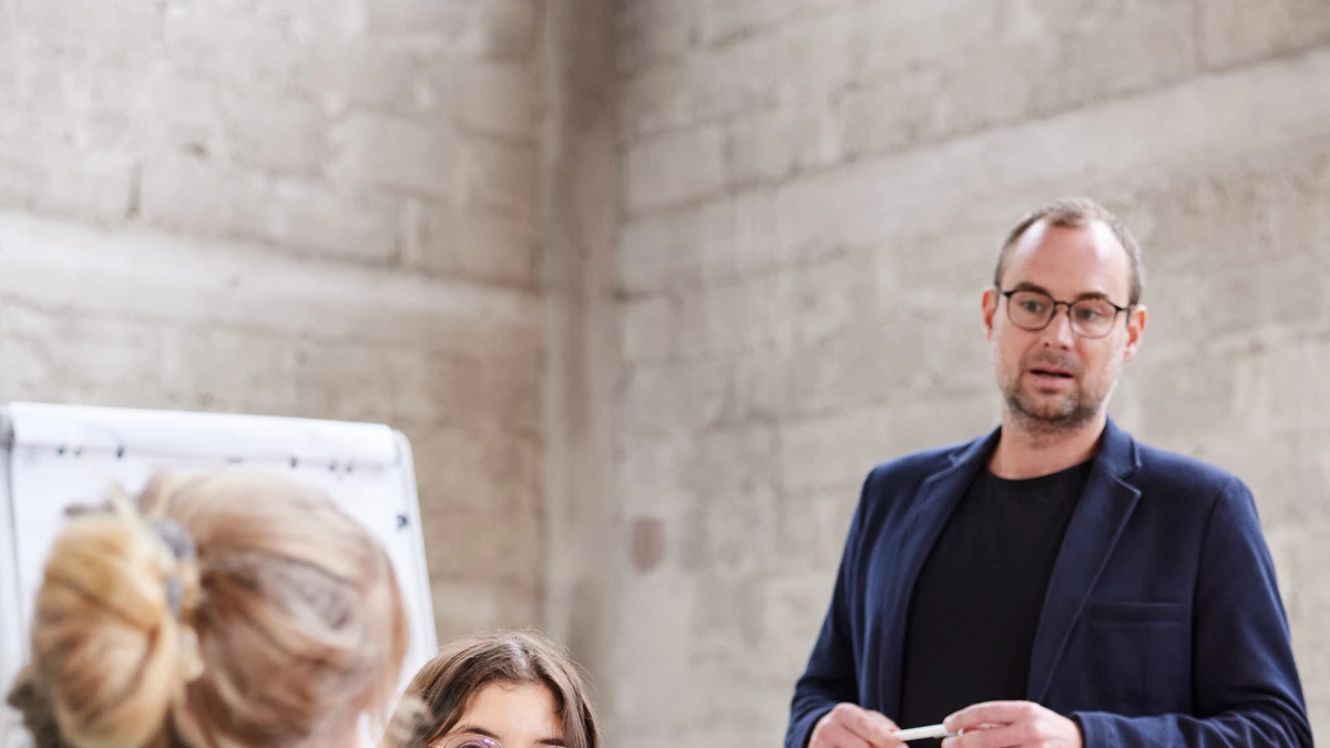 Das Bild zeigt drei Personen in einem Besprechungsraum. Eine Person, ein Mann mit Brille, steht im Hintergrund neben einem Laptop und hält einen Stift. Im Vordergrund sitzen zwei Frauen an einem Tisch. Die Frau links im türkisfarbenen Blazer spricht gestikulierend mit der Frau gegenüber, die ein beiges Oberteil und eine bunte Schal trägt. Auf dem Tisch liegen einige Papiere, Stifte und Wassergläser. Im Hintergrund ist eine Betonwand und ein Flipchart zu sehen.