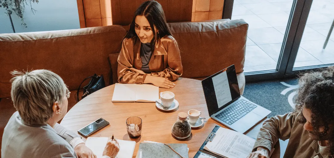 Selbstpräsentation im Vorstellungsgespräch: Drei Personen sitzen in entspannter Atmosphäre am Tisch, eine Bewerberin stellt sich vor, während zwei Recruiter Notizen machen und Unterlagen prüfen.