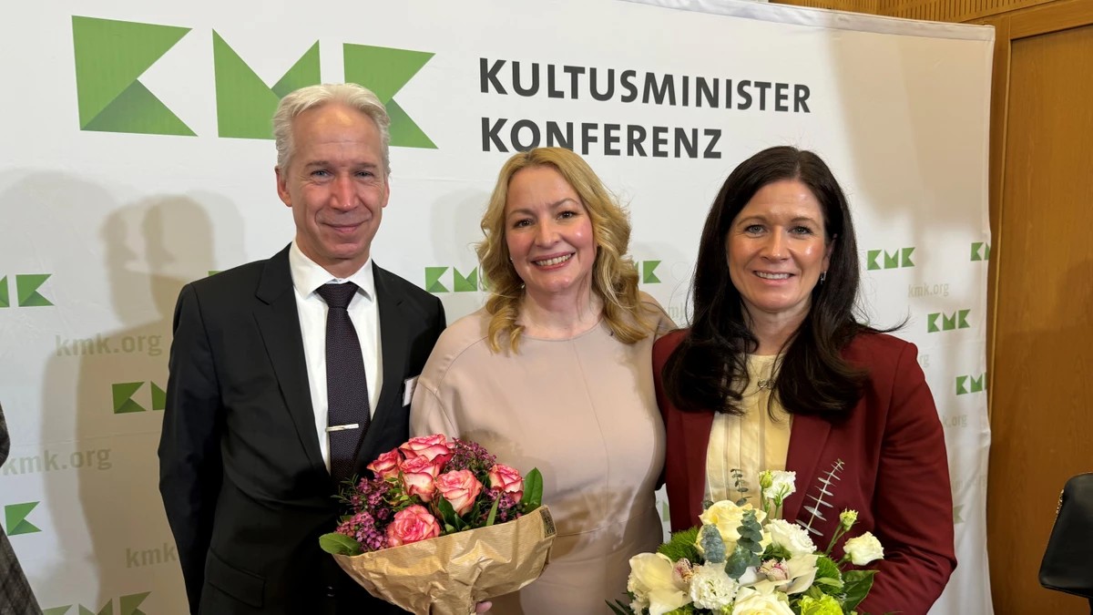 Es sind drei Personen vor der Fotowand der Kultusministerkonferenz zu sehen. Die beiden Frauen haben Blumensträuße in der Hand. Ihnen wird zur Verabschiedung bzw. zur Neuwahl gratuliert.