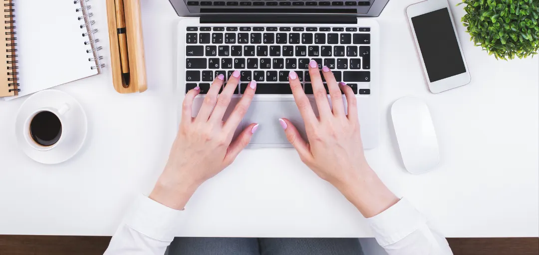 Arbeitszeugnis auf dem Laptop lesen: Hände tippen auf der Tastatur neben Kaffee und Notizbuch. 