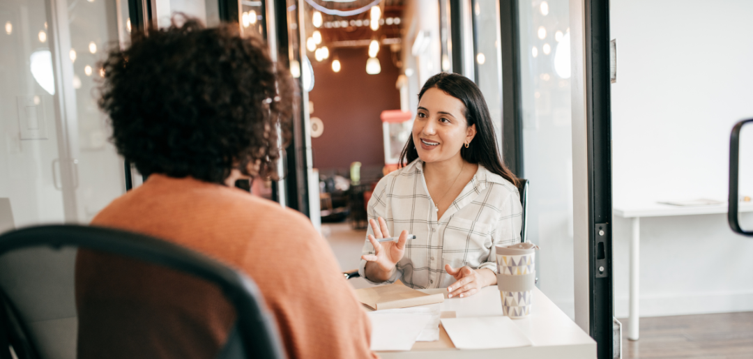 Zwei Frauen im Bewerbungsgespräch. Die Interviewerin sitzt mit dem Rücken zur Kamera. Die Bewerberin erklärt ihr lächelnd etwas. 