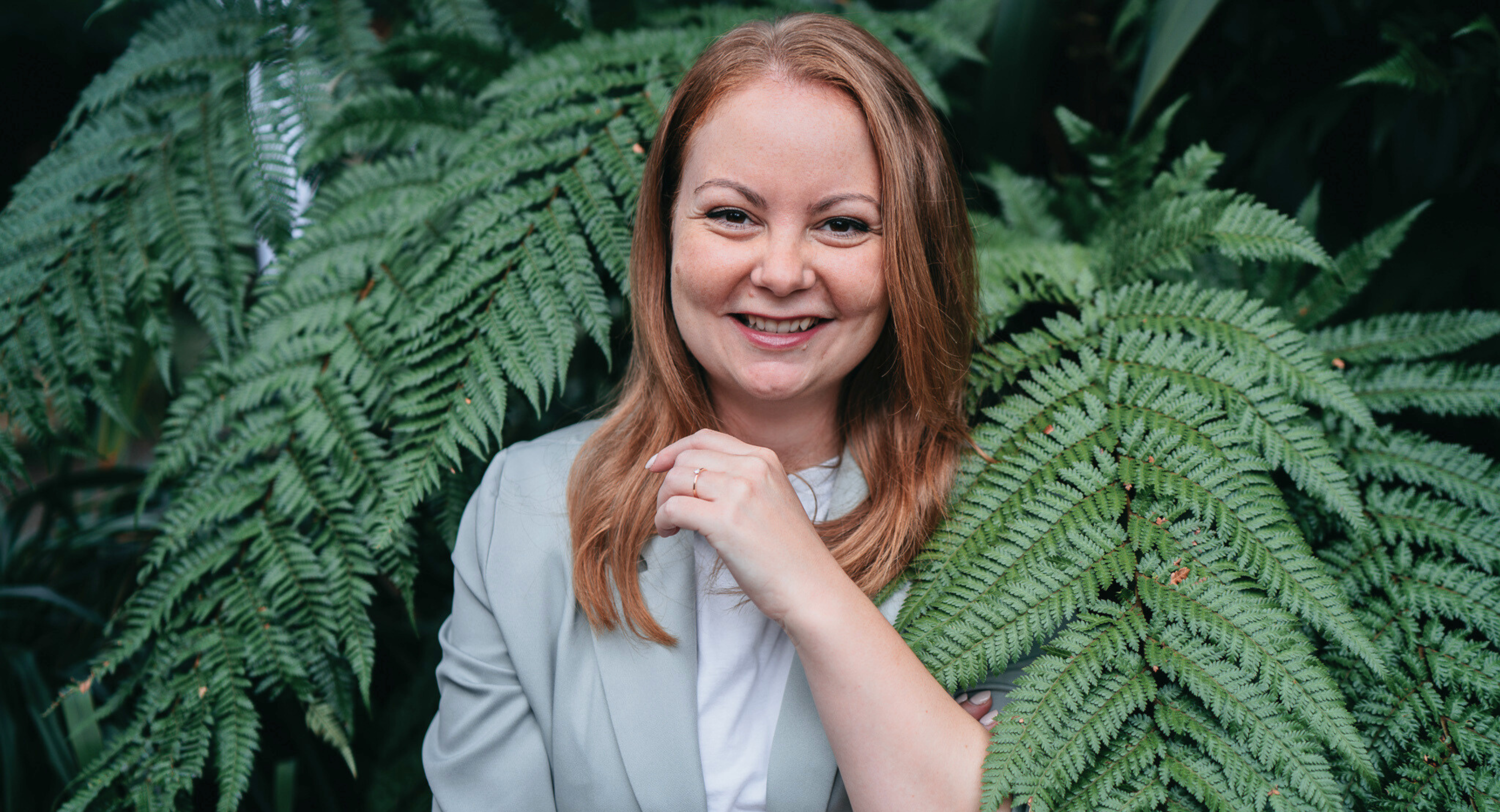 Vanessa Danesan steht lächelnd in weißer Bluse und grauem Blazer vor einem Farn. 