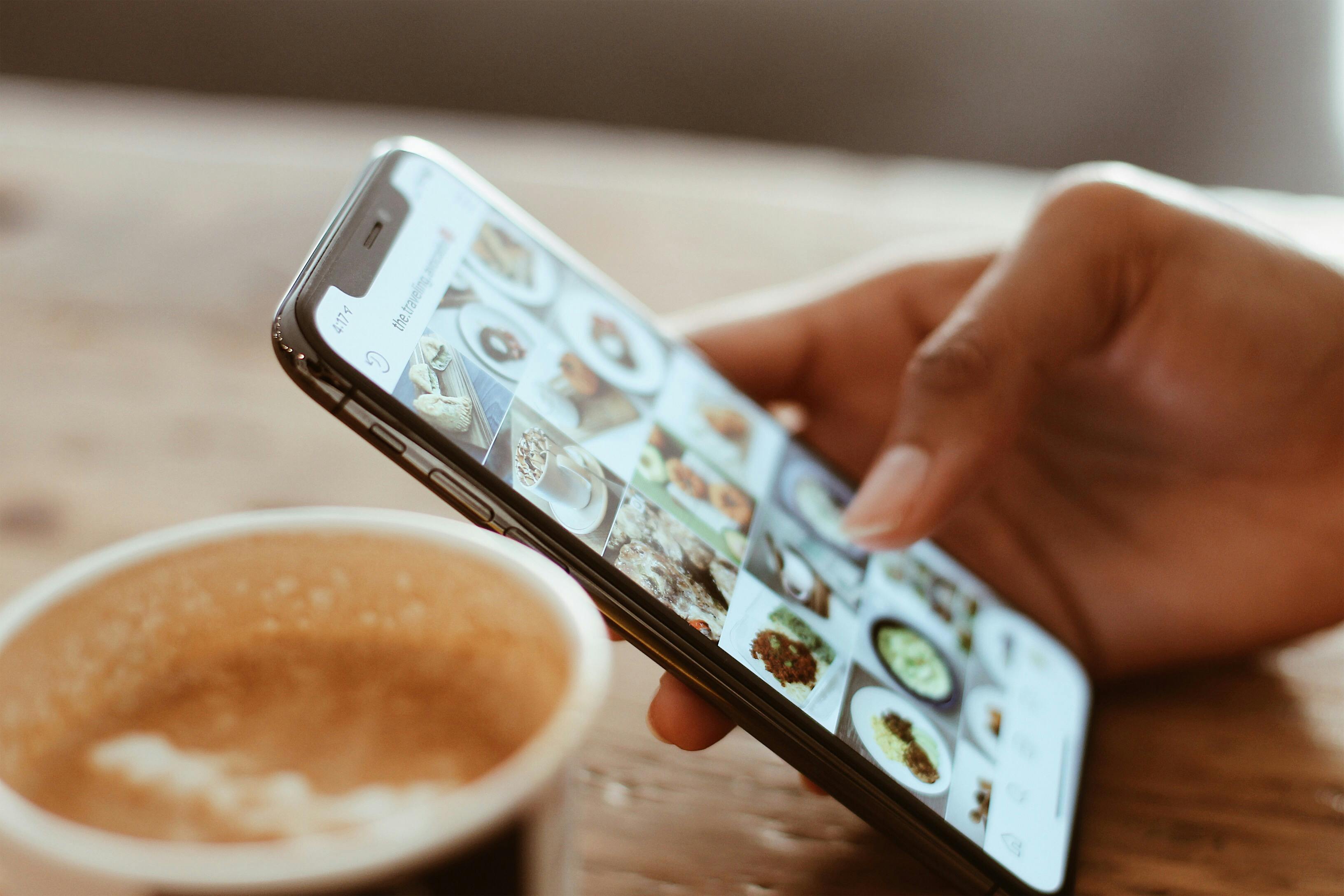 Ein Smartphone, auf dem eine Bild-Feed-App geöffnet ist. Daneben steht ein Cappucino im To-Go-Becher.