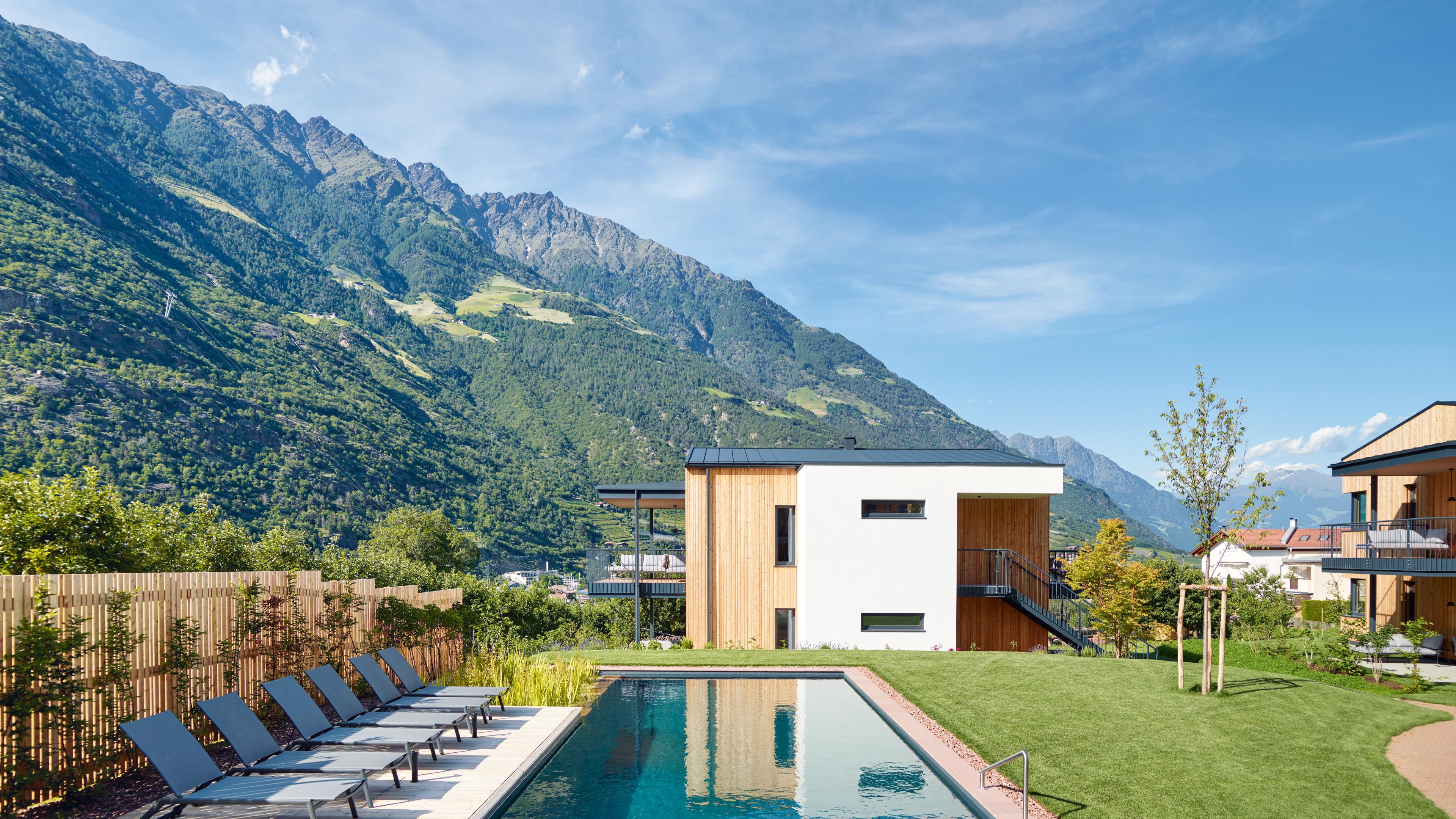 Über einen Pool und Badeliegen blickt man auf eine moderne Holzlodge vor Bergpanorama. 