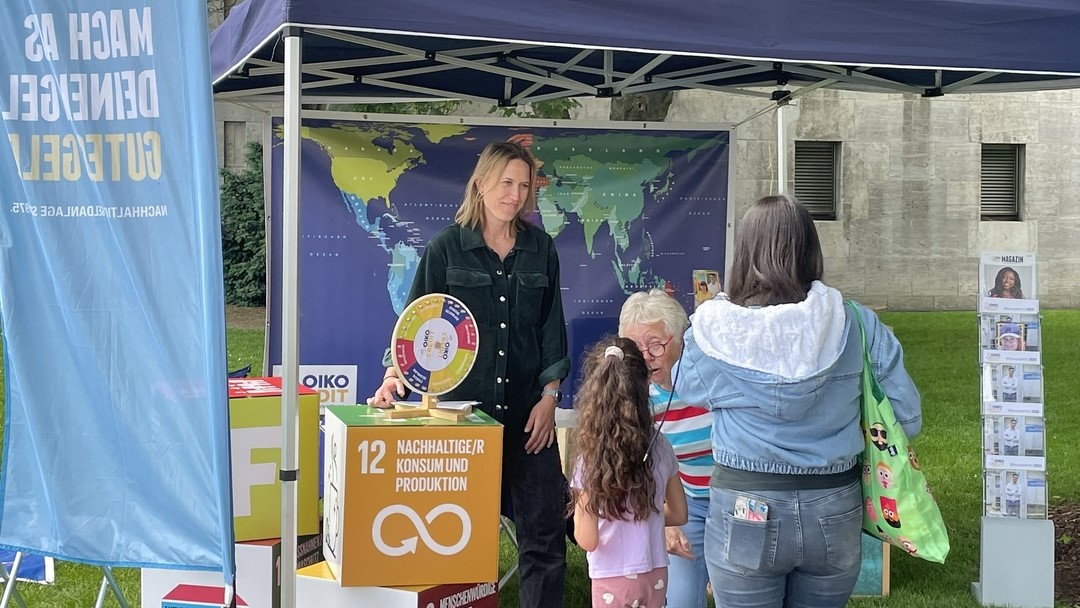 Eine Frau steht unter einem Pavillon am Stand von Oikocredit. an dem es ein Glücksrad gibt. Den Stand besuchen eine Frau und ein junges Mädchen, die sich mit einer weiteren Frau unterhalten. 